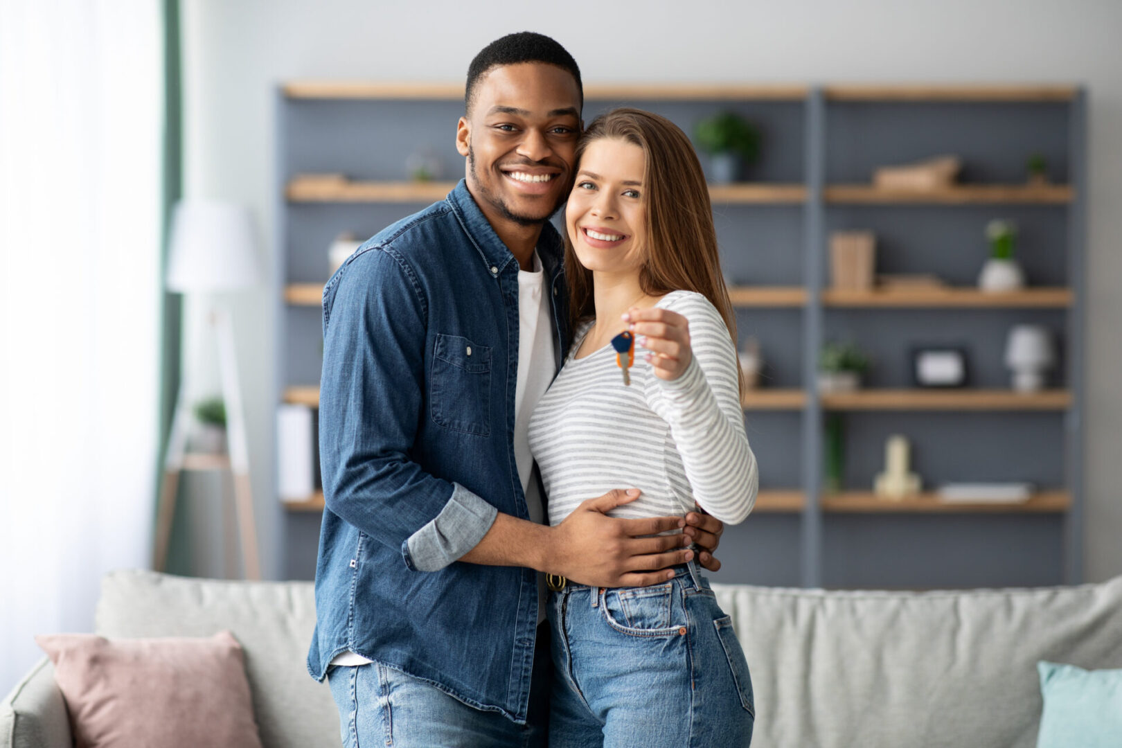 Happy young multiracial couple holding home keys and hugging, romantic millennial family, black man and white woman poising in living room after moving to new appartment, smiling at camera, free space