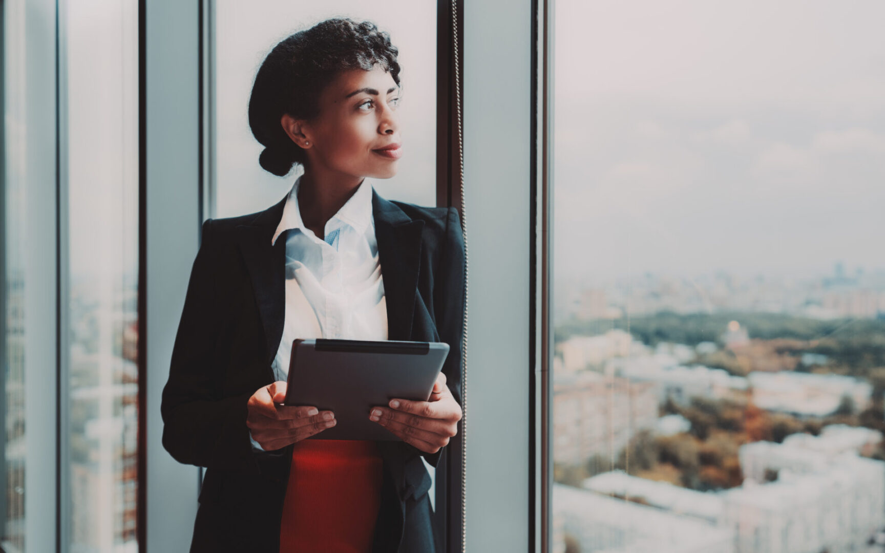 A dazzling young African-American woman entrepreneur with a digital tablet in her hands is standing near a huge panoramic window indoors of a business high-rise and pensively looking at the cityscape