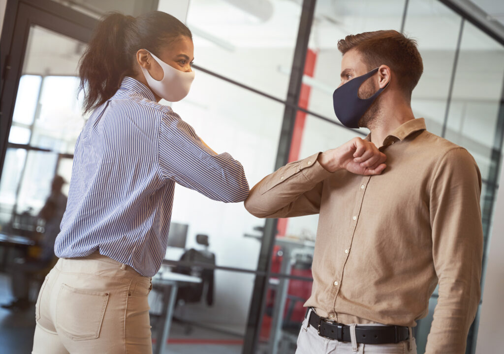 Social distancing at work. Two young diverse business colleagues wearing face protective masks bumping elbows, greeting each other while working during covid 19 quarantine. Preventing coronavirus